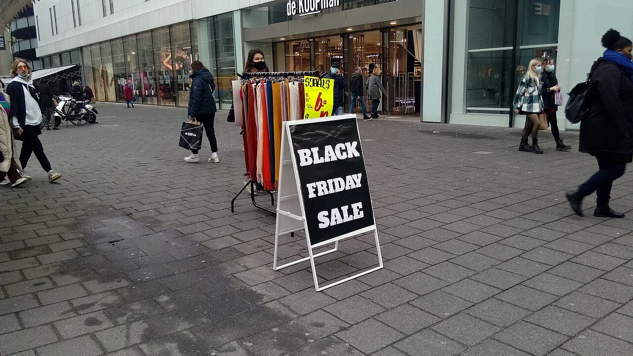 Placa de calçada com os dizeres BLACK FRIDAY SALE em frente a uma loja, com araras de roupas e pessoas circulando na rua comercial