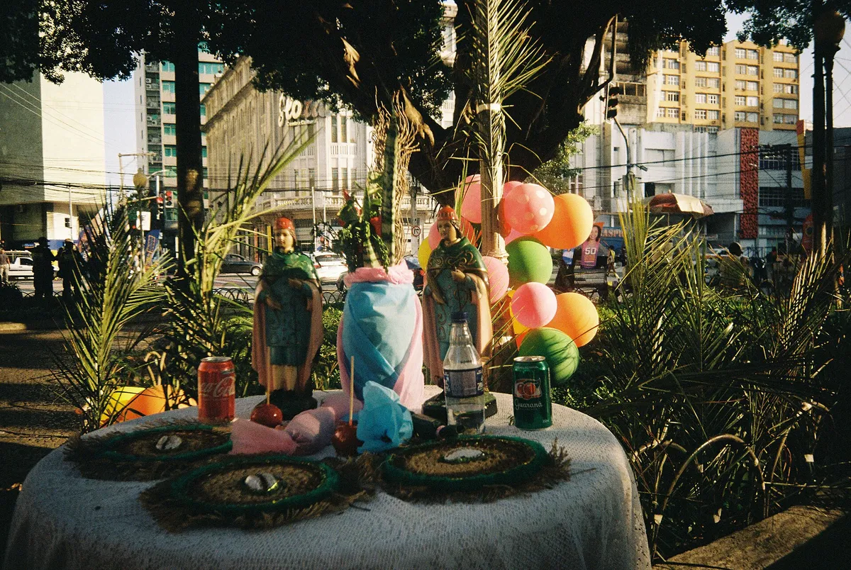 Altar ao ar livre com imagens de Cosme e Damião sobre uma mesa, cercadas de balões coloridos, latas de refrigerante e doces