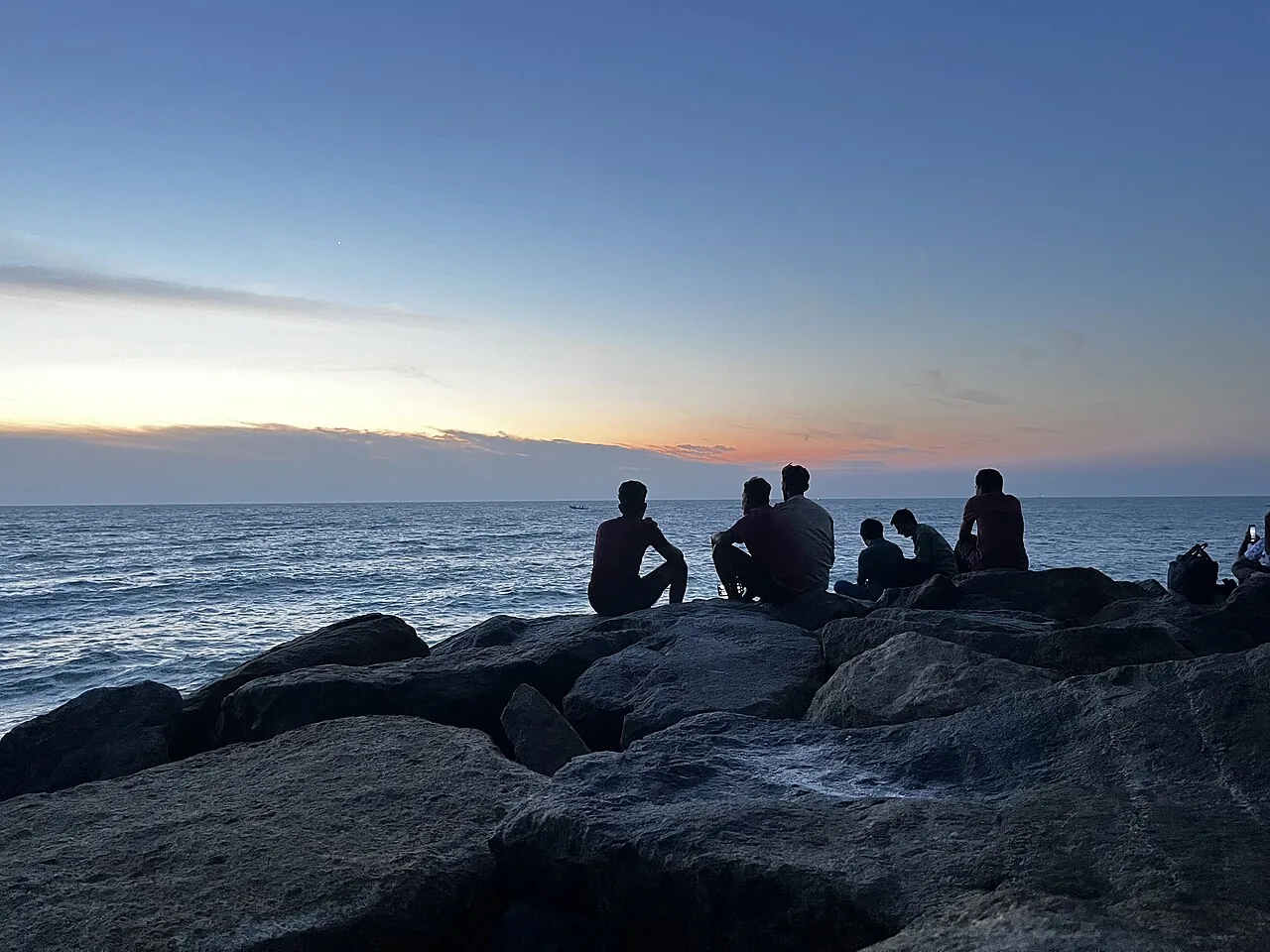 Grupo de amigos sentado lado a lado sobre pedras assistindo ao pôr do sol no mar, no Dia do Amigo