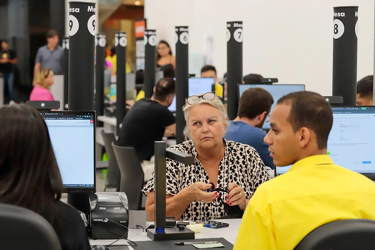 Salão de atendimento ao público com mesas numeradas e servidores atendendo cidadãos em computadores