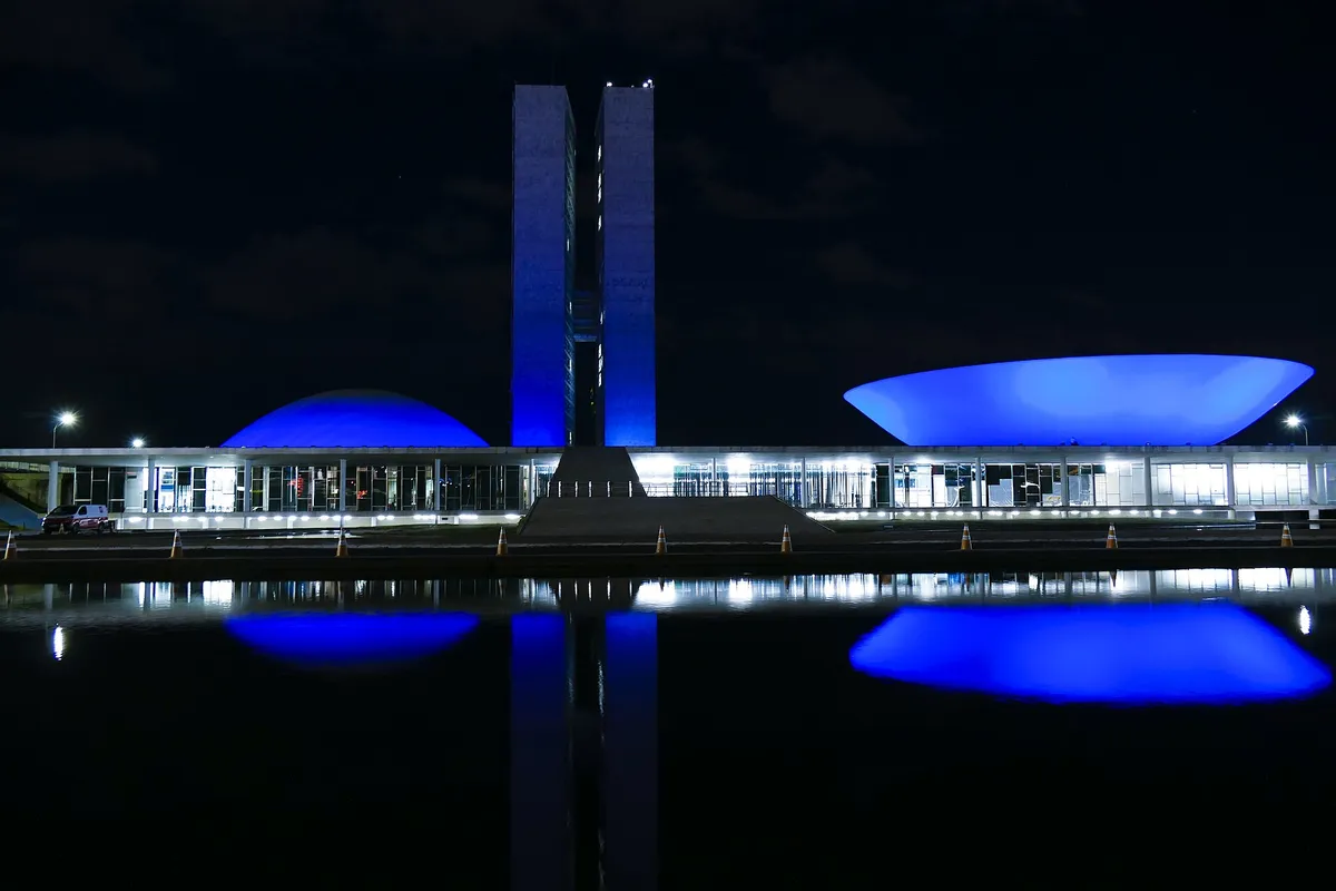 Prédio do Congresso Nacional à noite, com as duas cúpulas e as torres iluminadas de azul e refletidas no espelho d'água