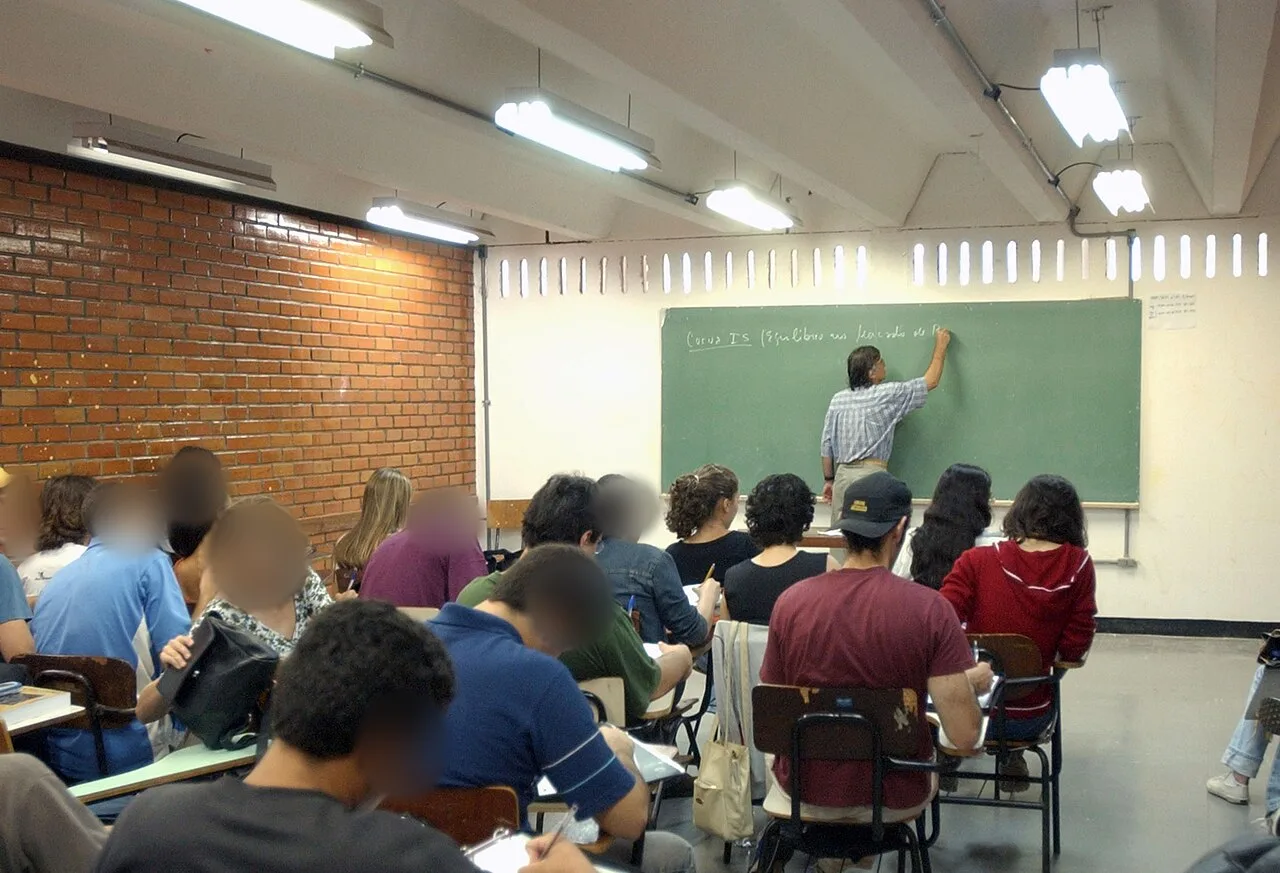 Estudantes de costas em sala de aula, com professor escrevendo no quadro verde, na preparação para o Enem