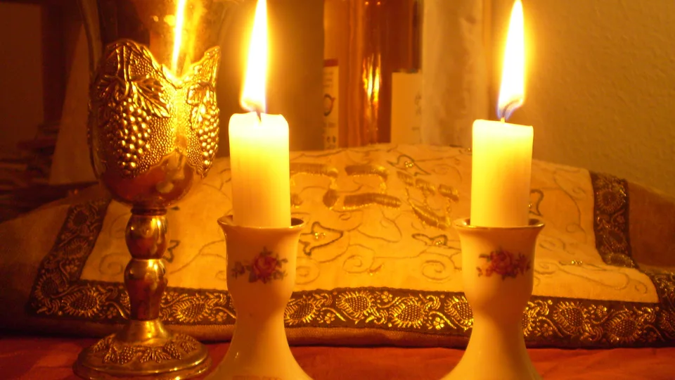 Duas velas acesas em castiçais e um cálice dourado sobre a mesa, como se faz na abertura do Shabat