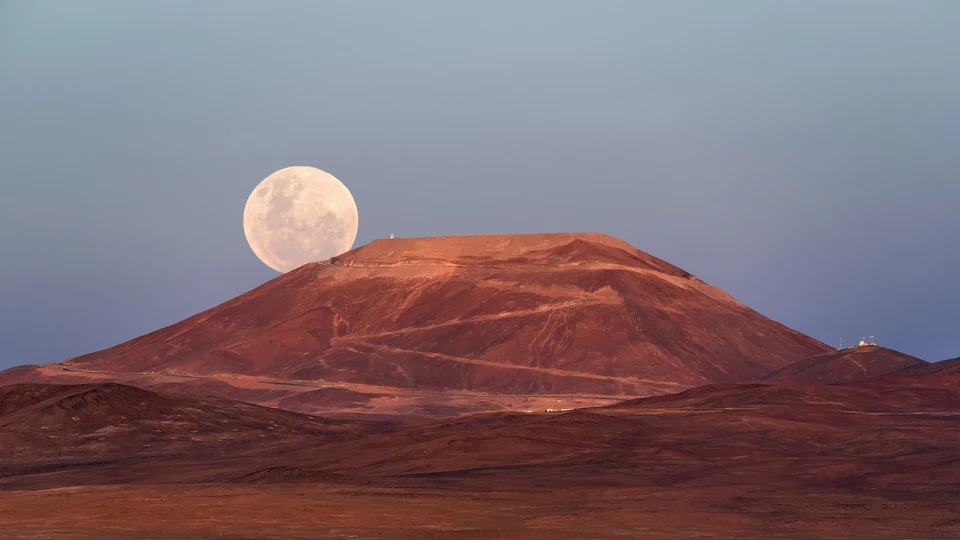 Superlua grande e clara nascendo atrás de uma montanha avermelhada no deserto, ao entardecer