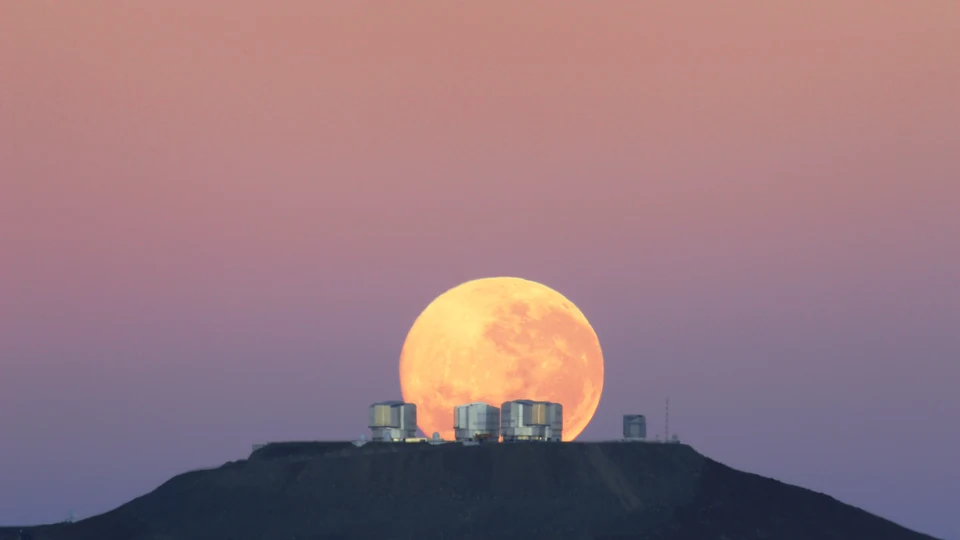 Lua cheia se pondo atrás de um observatório no alto de uma montanha, com o céu rosado ao amanhecer