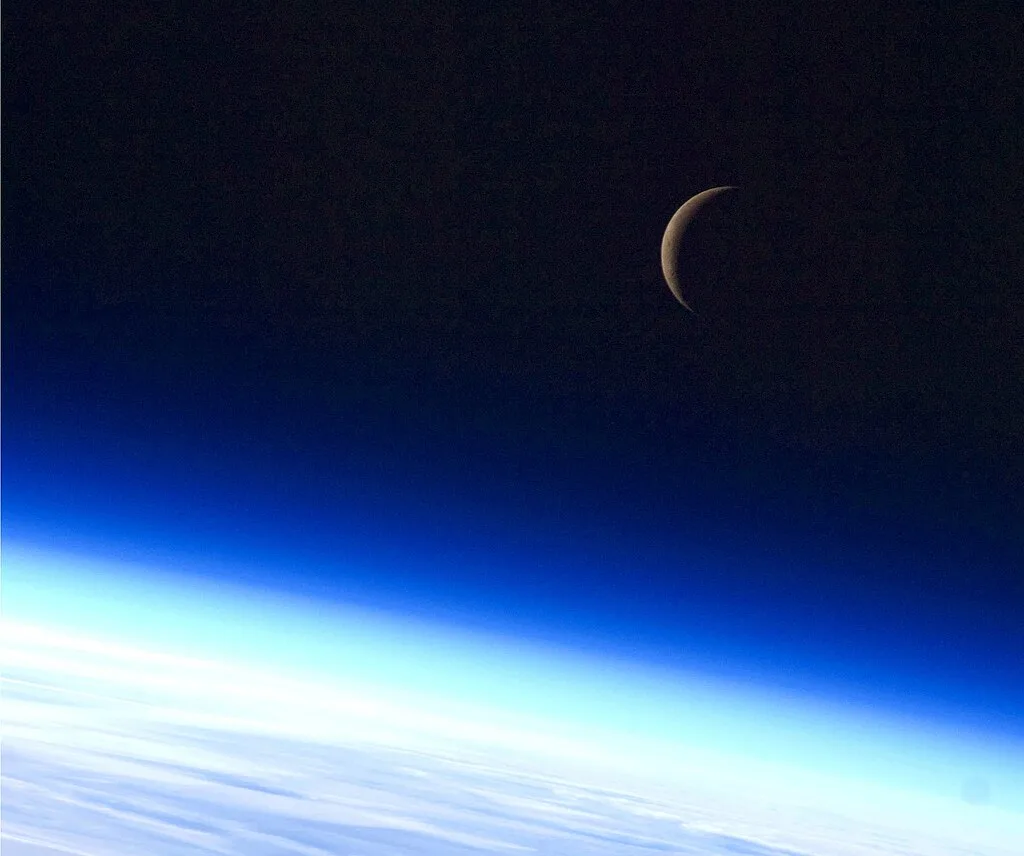 Lua crescente vista do espaço pela tripulação da Estação Espacial Internacional, com a curvatura da atmosfera terrestre ao fundo.
